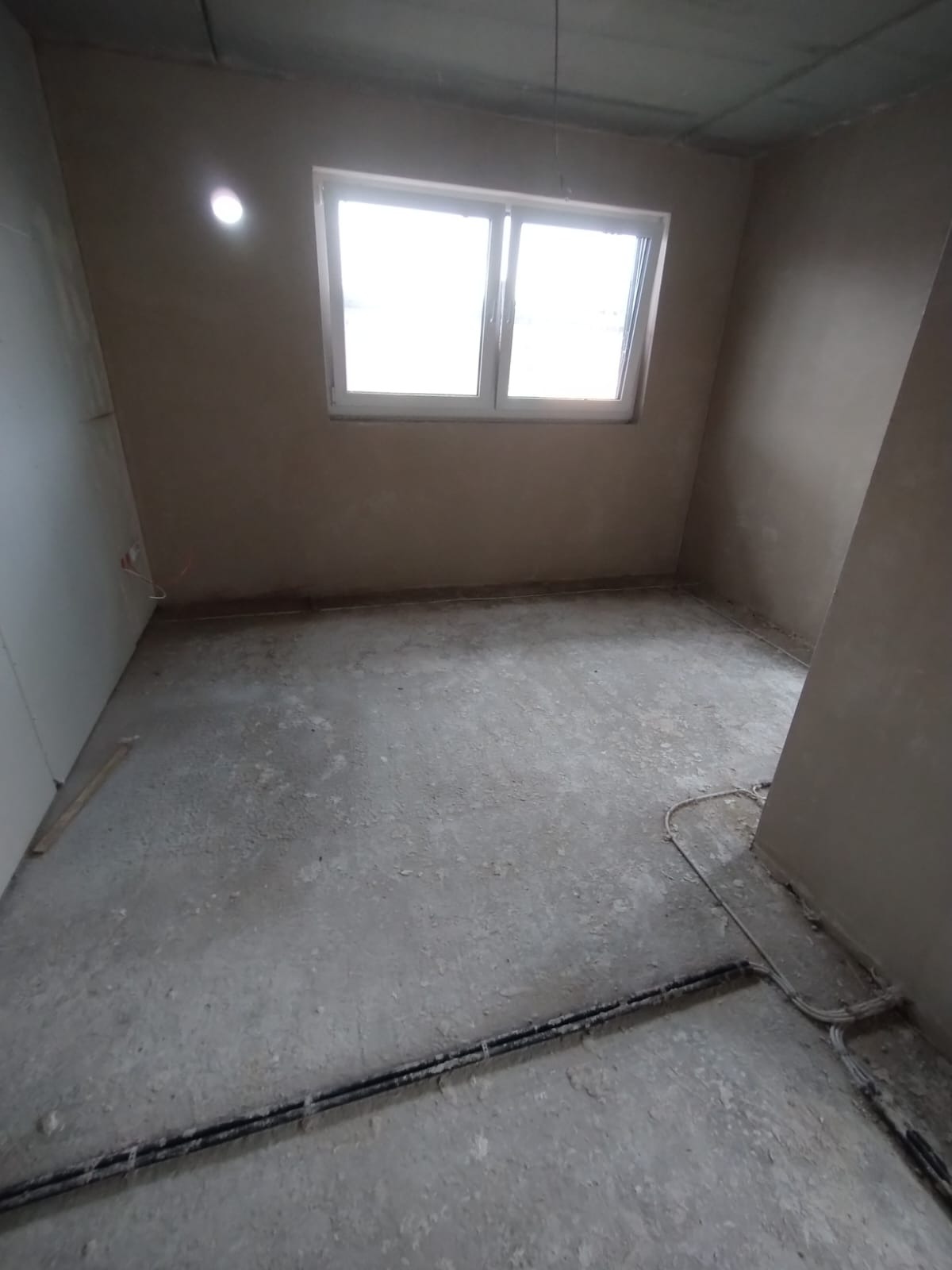 Pokój po nałożeniu tynku, widoczne ściany w szarym odcieniu, okno z białymi ramami i instalacja elektryczna przy podłodze. Surowy stan wykończenia wnętrza.