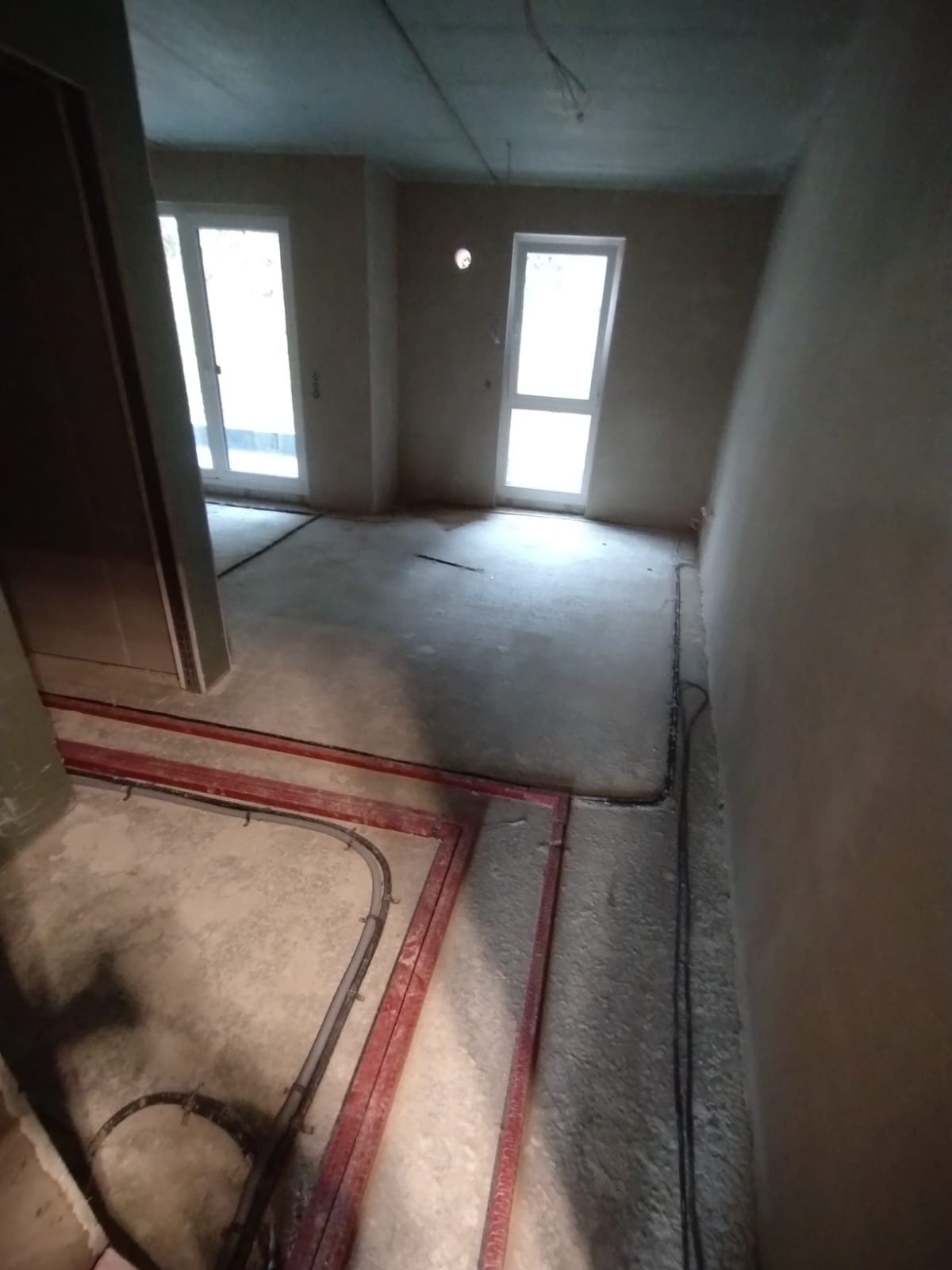 Surowy stan wnętrza z instalacją ogrzewania podłogowego w czerwonej ramie i przewodami elektrycznymi przy ścianie, przygotowany do tynkowania lub szpachlowania.