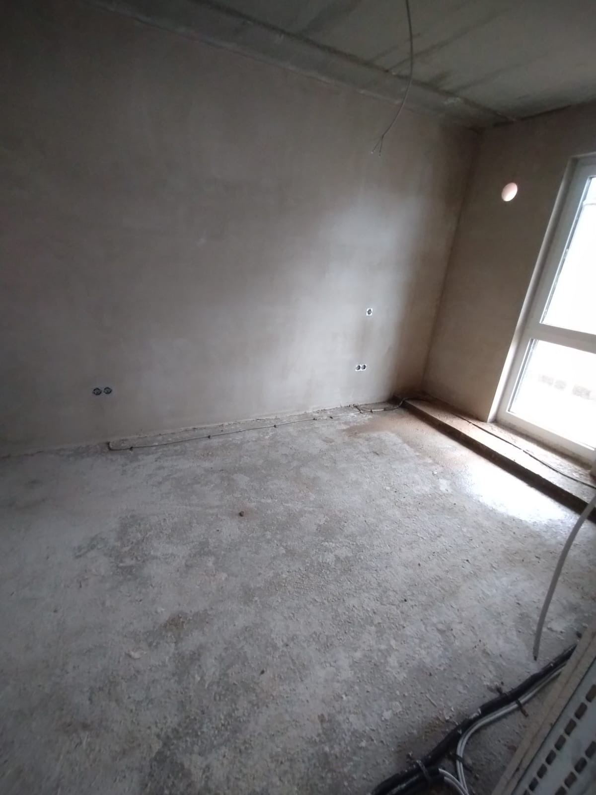Surowe wnętrze pokoju po tynkowaniu ścian, widoczne gniazdka elektryczne i okno z białymi ramami. Podłoga betonowa, przygotowana do dalszych prac wykończeniowych.