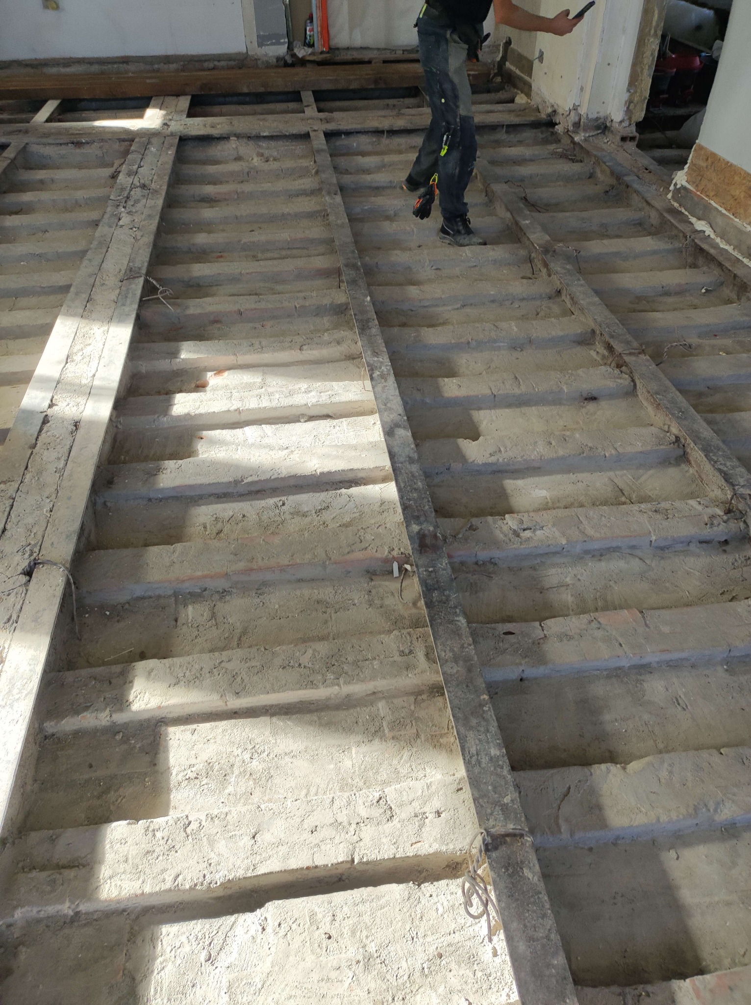 Demontaż podłogi: odsłonięte legary i ceglane wypełnienie w trakcie prac rozbiórkowych. Widoczny fragment osoby z telefonem. Oświetlenie naturalne.