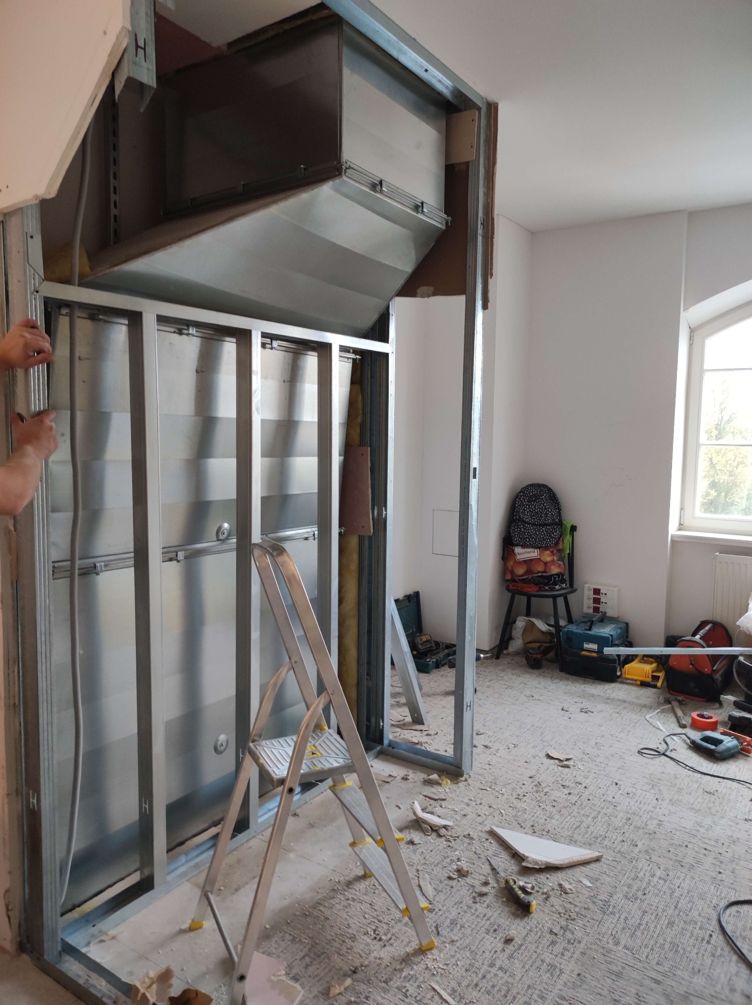 Montaż ścianki działowej z metalowych profili w jasnym pomieszczeniu. Widoczna drabina, narzędzia i elementy instalacji wentylacyjnej. Częściowy widok osoby po lewej stronie.