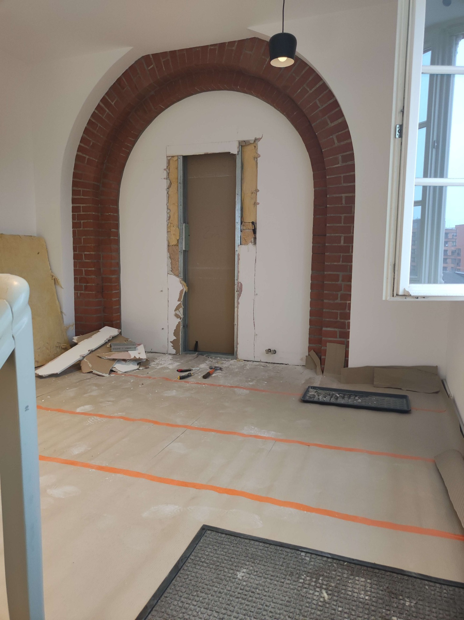 Wnętrze w trakcie prac: wyburzona ściana z ceglanym łukiem, widoczne narzędzia i gruz. Podłoga zabezpieczona folią. Okno z widokiem na budynki.
