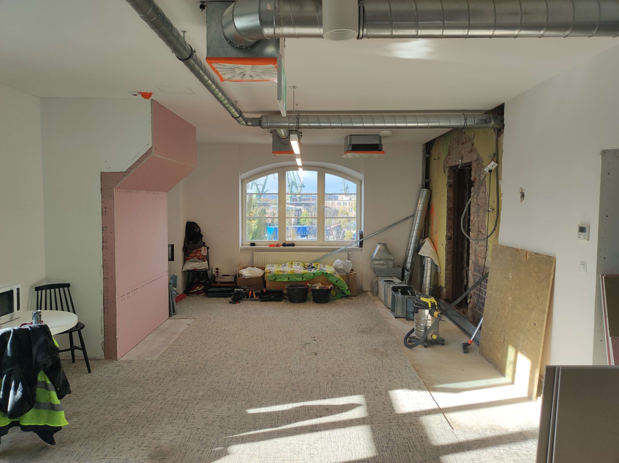 Wnętrze w trakcie remontu: odsłonięta cegła, instalacje wentylacyjne, narzędzia, płyty gipsowe. Widok na okno z widokiem na miasto. Dziennie światło.