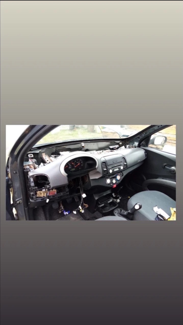 Wnętrze rozebranego samochodu marki Nissan Micra z widoczną deską rozdzielczą w trakcie naprawy lub demontażu. Widoczne kable i elementy elektroniki.