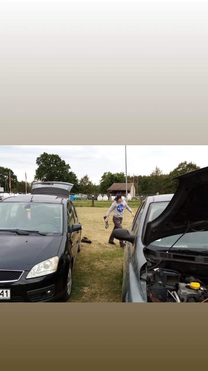 Dwa samochody z otwartymi maskami na trawie, osoba w bluzie NASA z narzędziem w dłoni przechodzi między nimi. W tle budynek i drzewa.