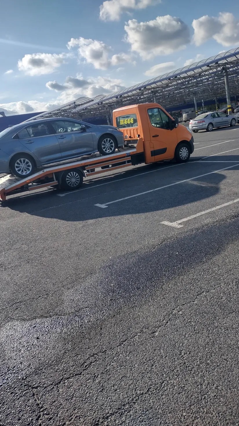 Pomarańczowa laweta z szarym samochodem osobowym na parkingu, widoczny numer telefonu. Transport pojazdu w słoneczny dzień.