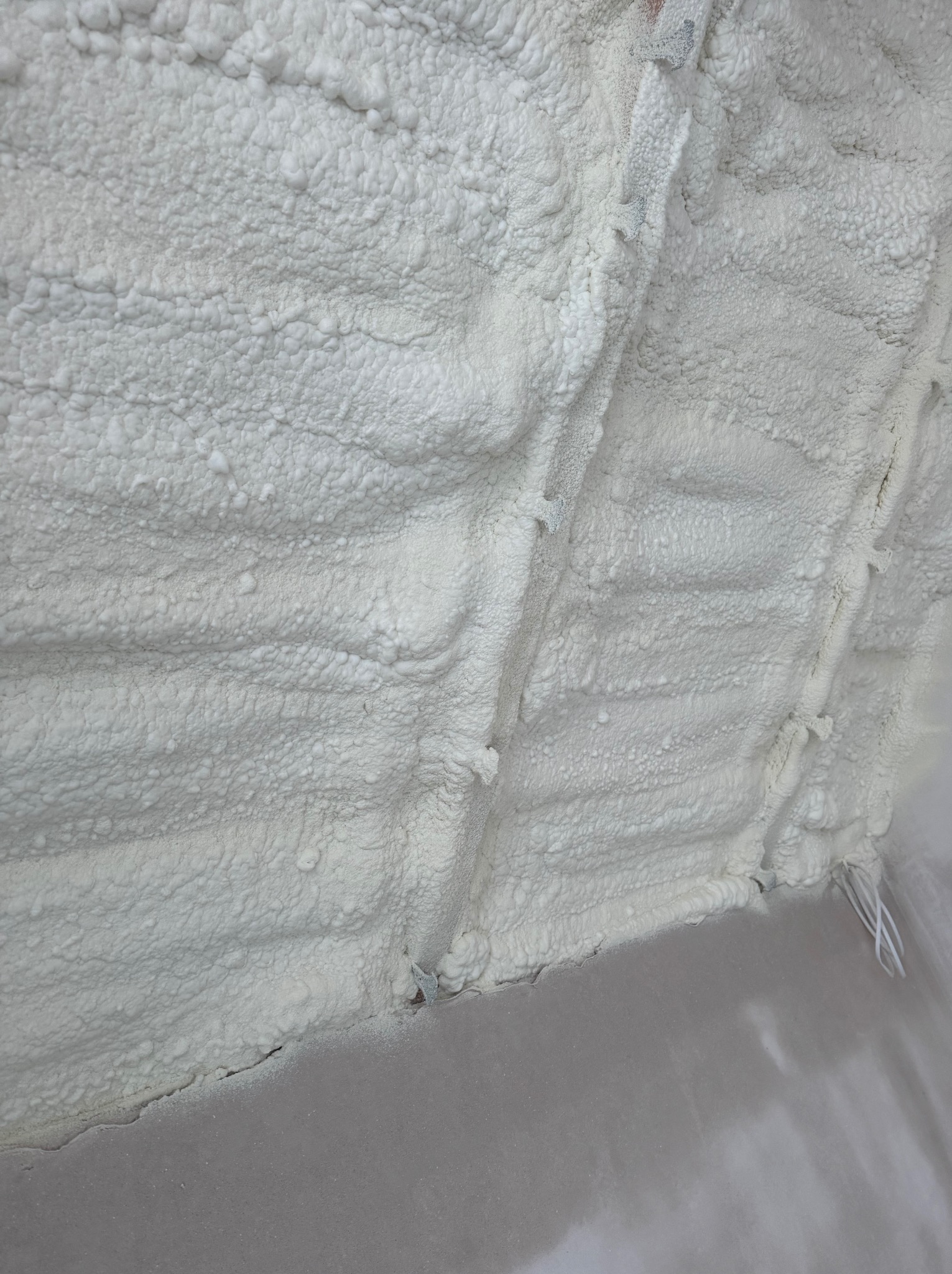 Szczegółowe ujęcie białej pianki izolacyjnej PUR, nierównomiernie nałożonej na powierzchnię, z widocznymi fragmentami podłoża w dolnej części kadru.