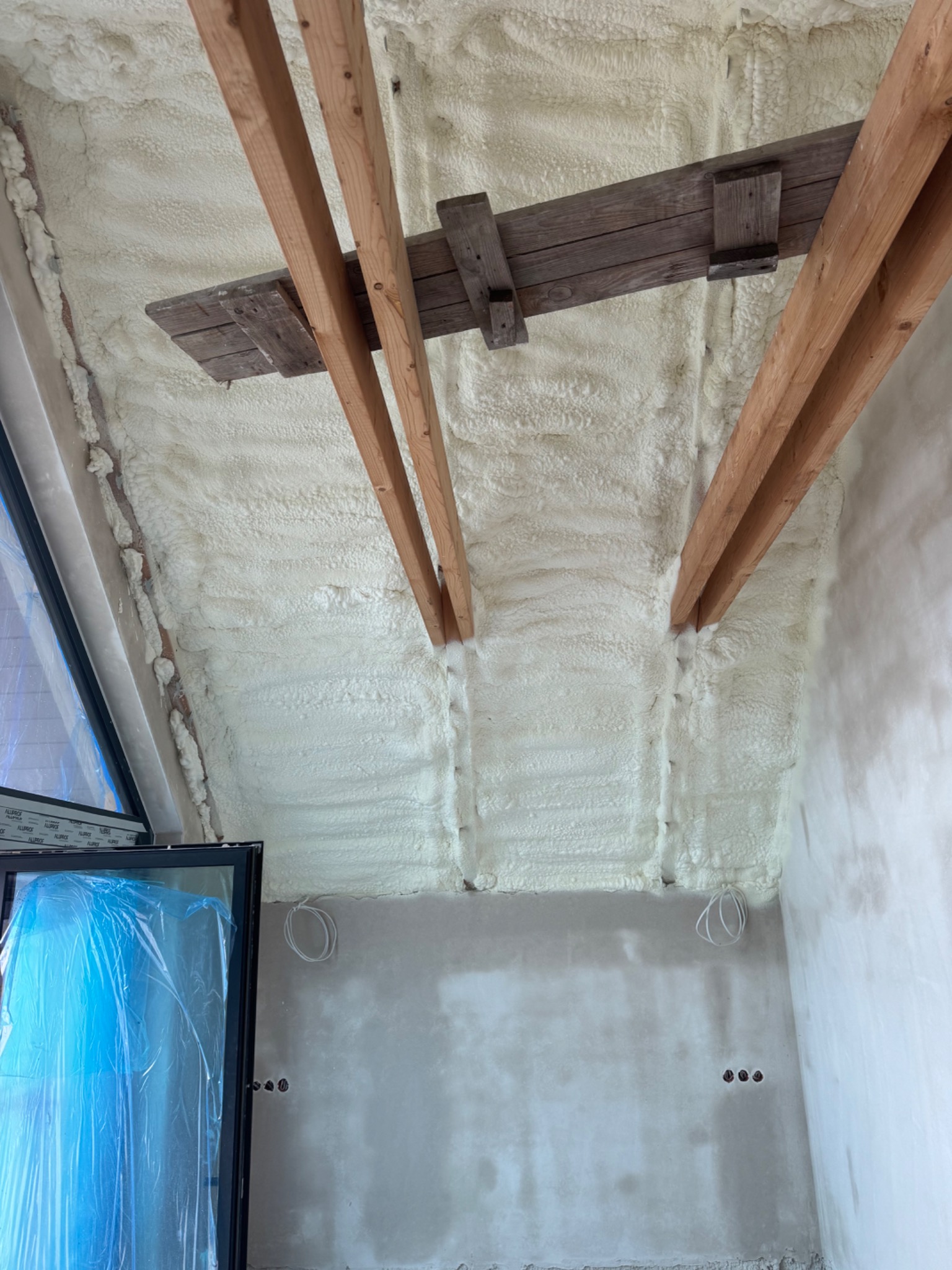 Poddasze po ociepleniu pianką PUR, widoczne drewniane belki konstrukcyjne i okno zabezpieczone folią. Ściany przygotowane do dalszych prac wykończeniowych.