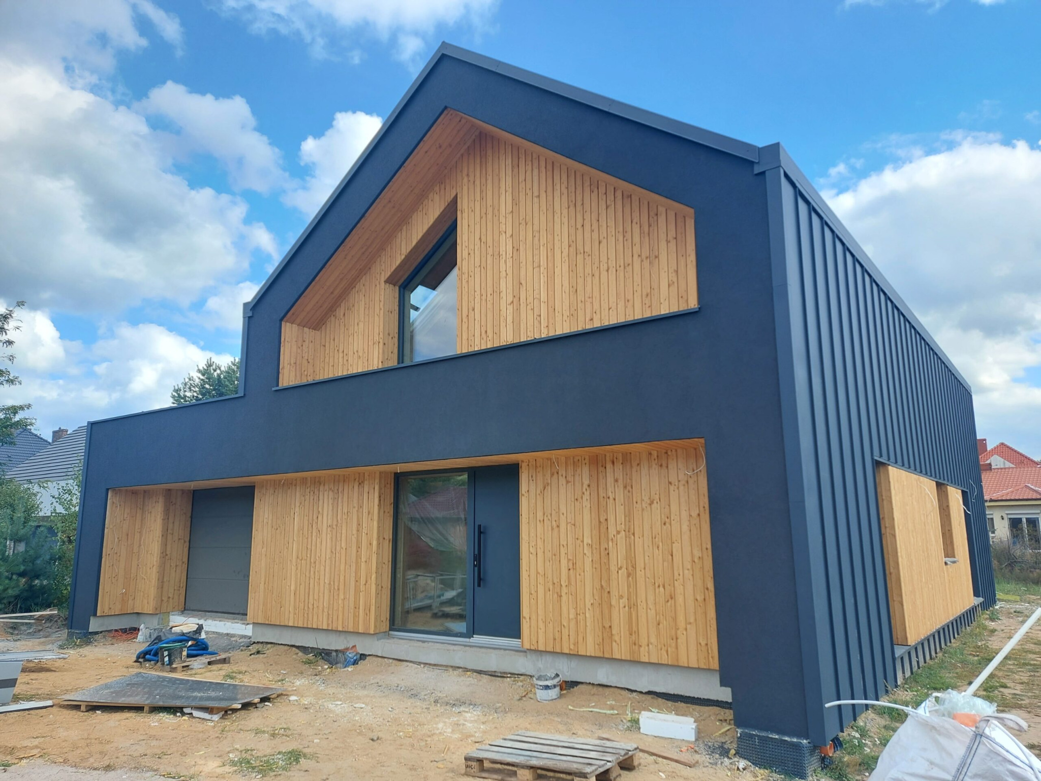 Nowoczesny dom z ciemną elewacją i drewnianą okładziną. Widoczne detale wykończenia, okna i drzwi. Tło stanowi błękitne niebo z chmurami.
