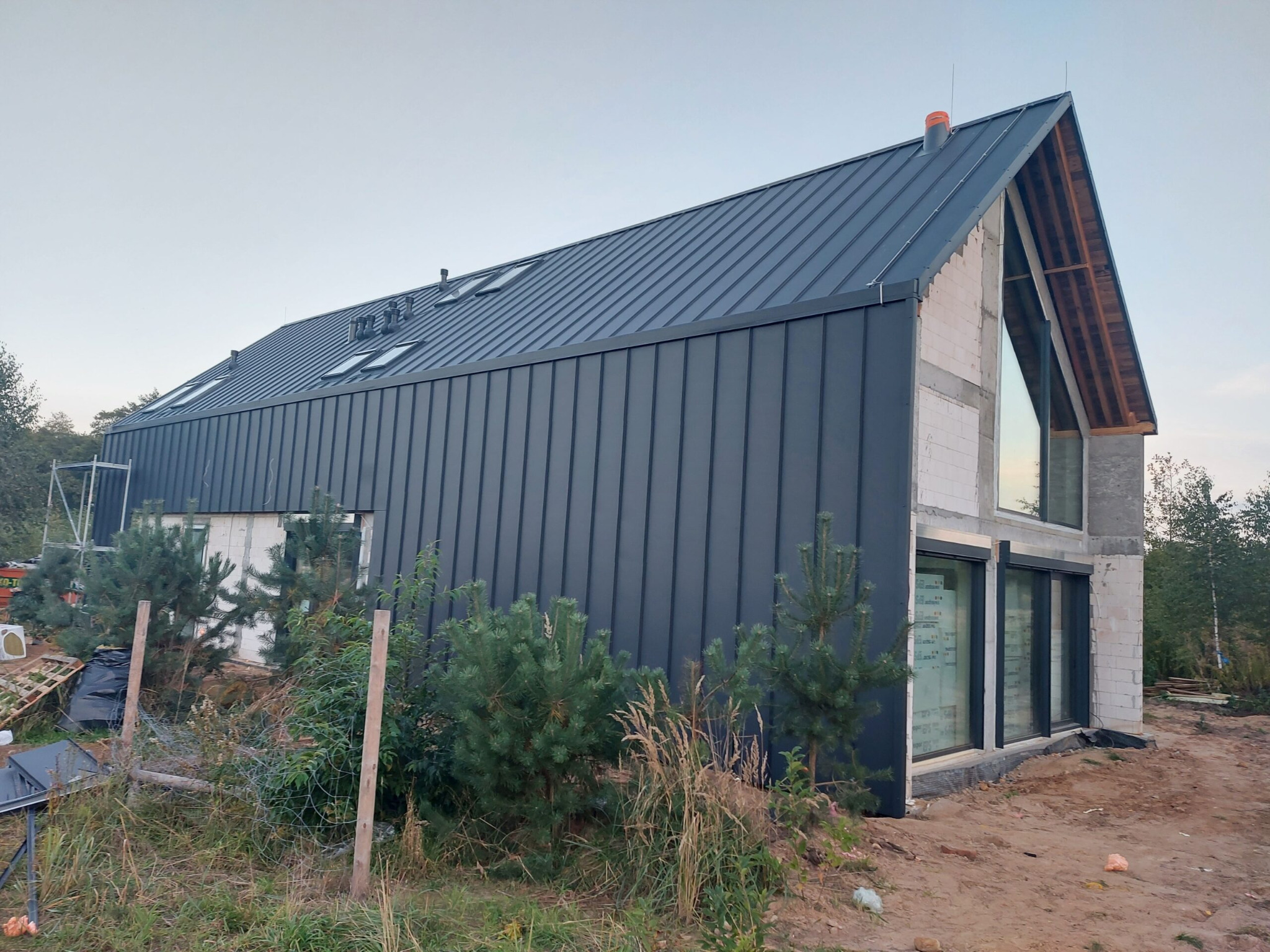 Nowoczesny dom w trakcie budowy z grafitową blachą na dachu i elewacji. Widoczne okna dachowe i duże przeszklenia. Surowy stan budynku, otoczenie z zielenią.