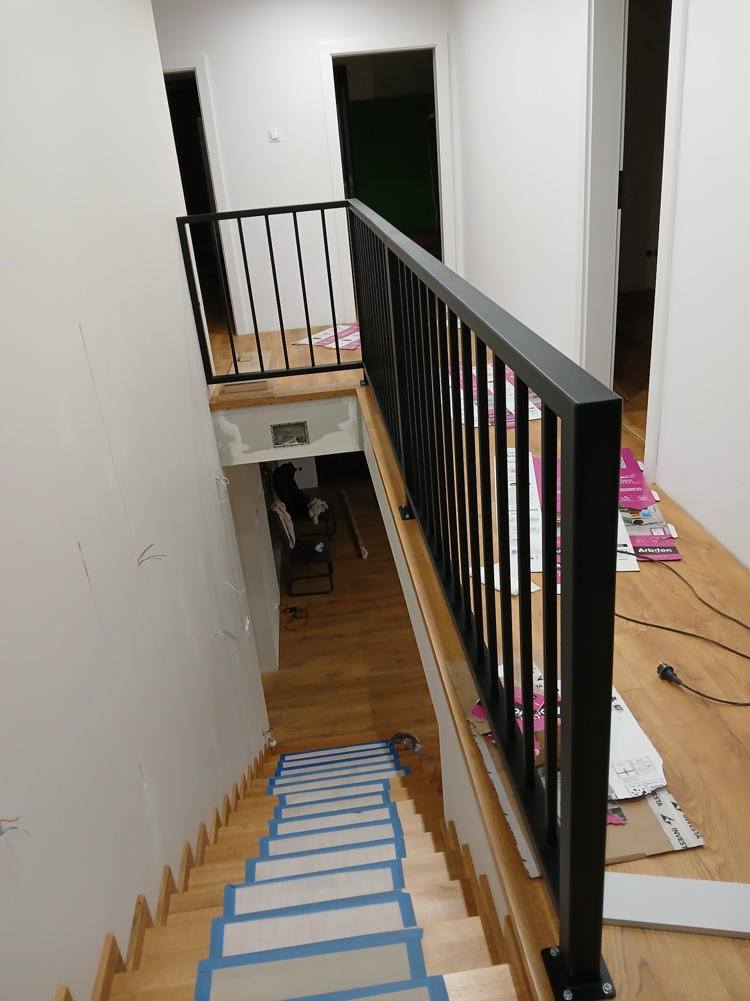 Nowoczesna, czarna balustrada schodowa z pionowymi szczeblami, zamontowana w jasnym wnętrzu. Schody zabezpieczone taśmą malarską, widoczne materiały budowlane.