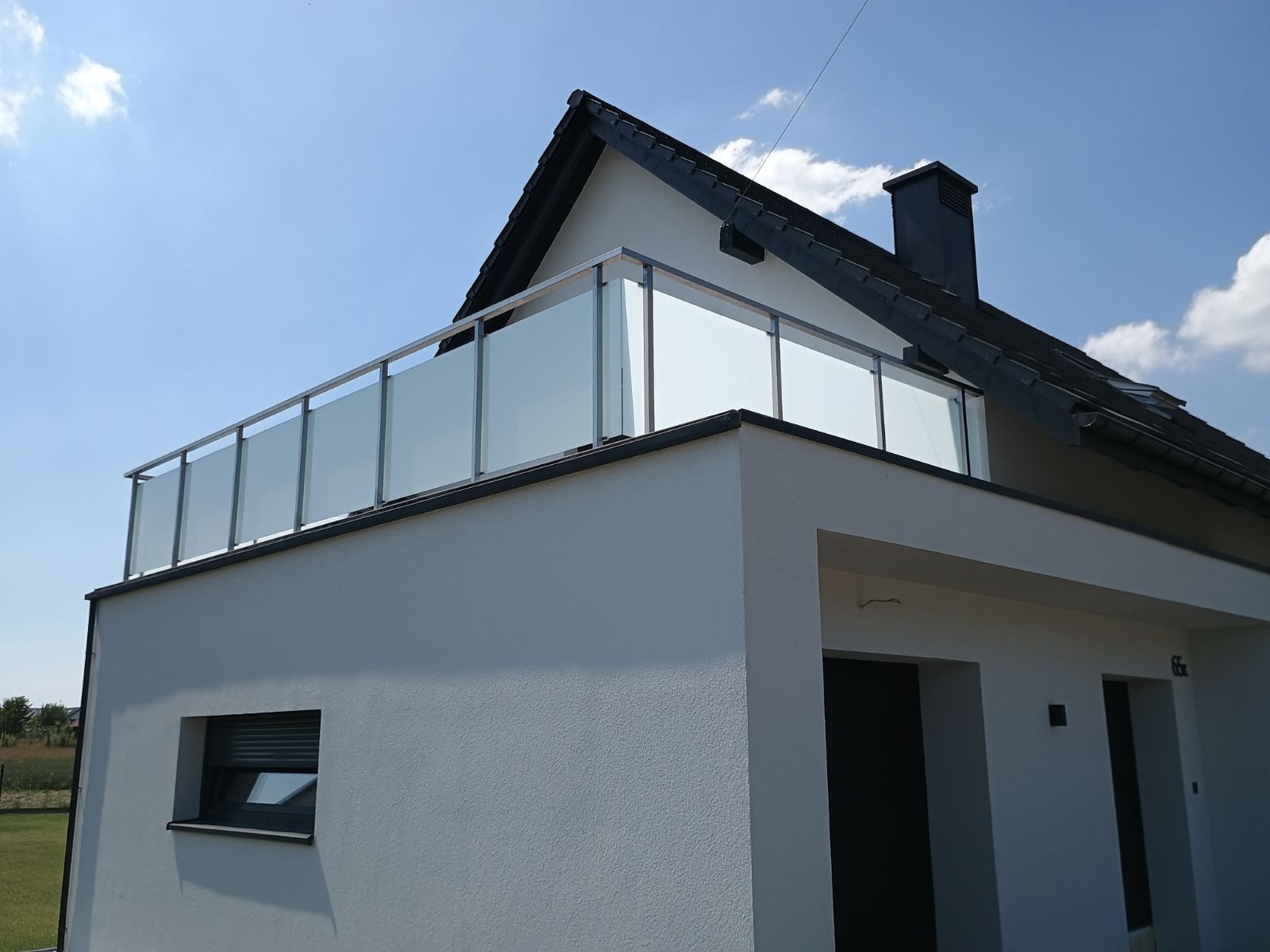 Nowoczesna, szklana balustrada balkonowa z metalowymi słupkami, zamontowana na białym budynku z ciemnym dachem, widziana z dołu pod kątem.