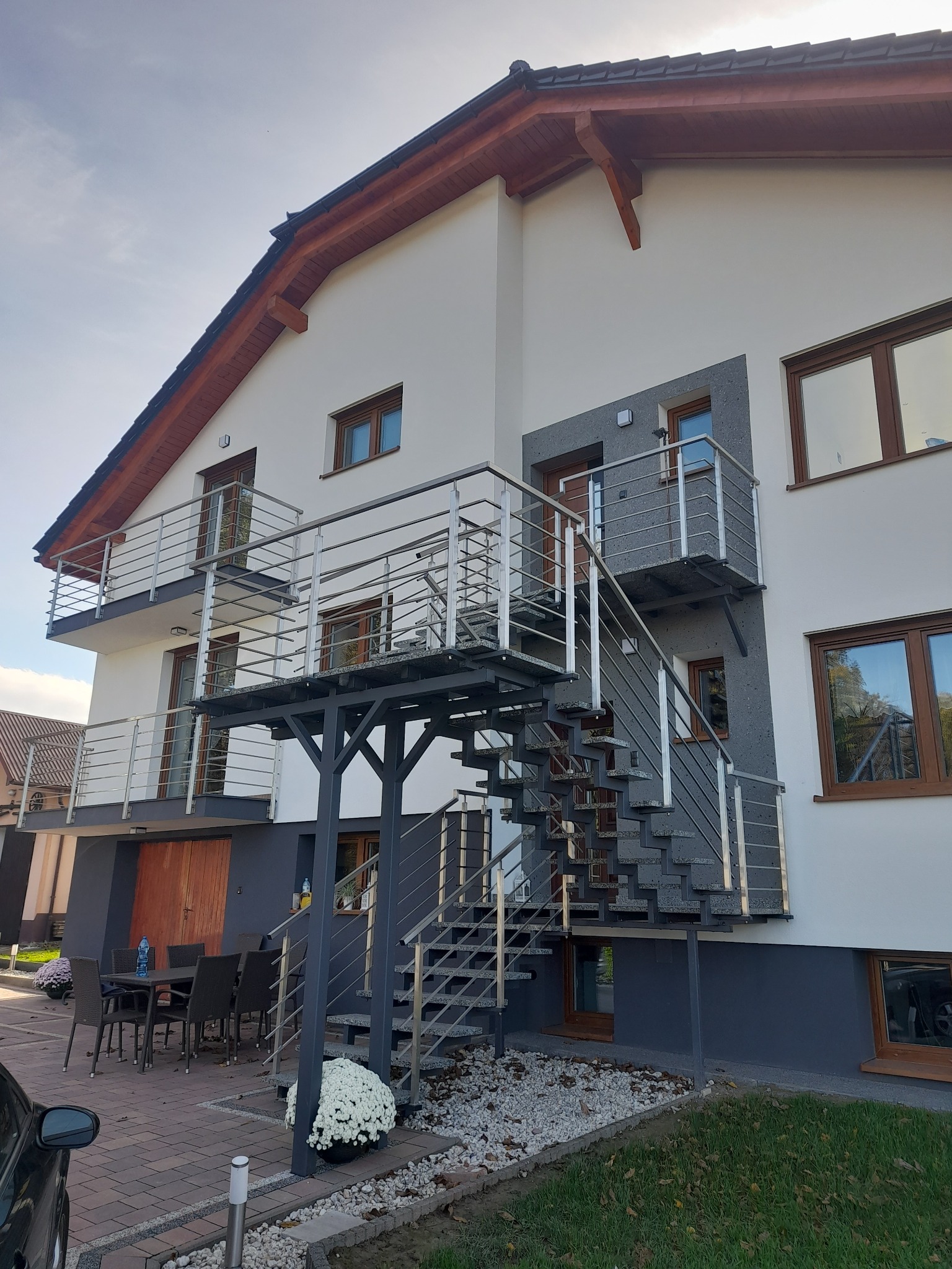 Dwukondygnacyjny budynek z zewnętrznymi schodami i balkonami z metalowymi barierkami. Schody prowadzą na górny balkon. Widoczne okna z drewnianymi ramami.