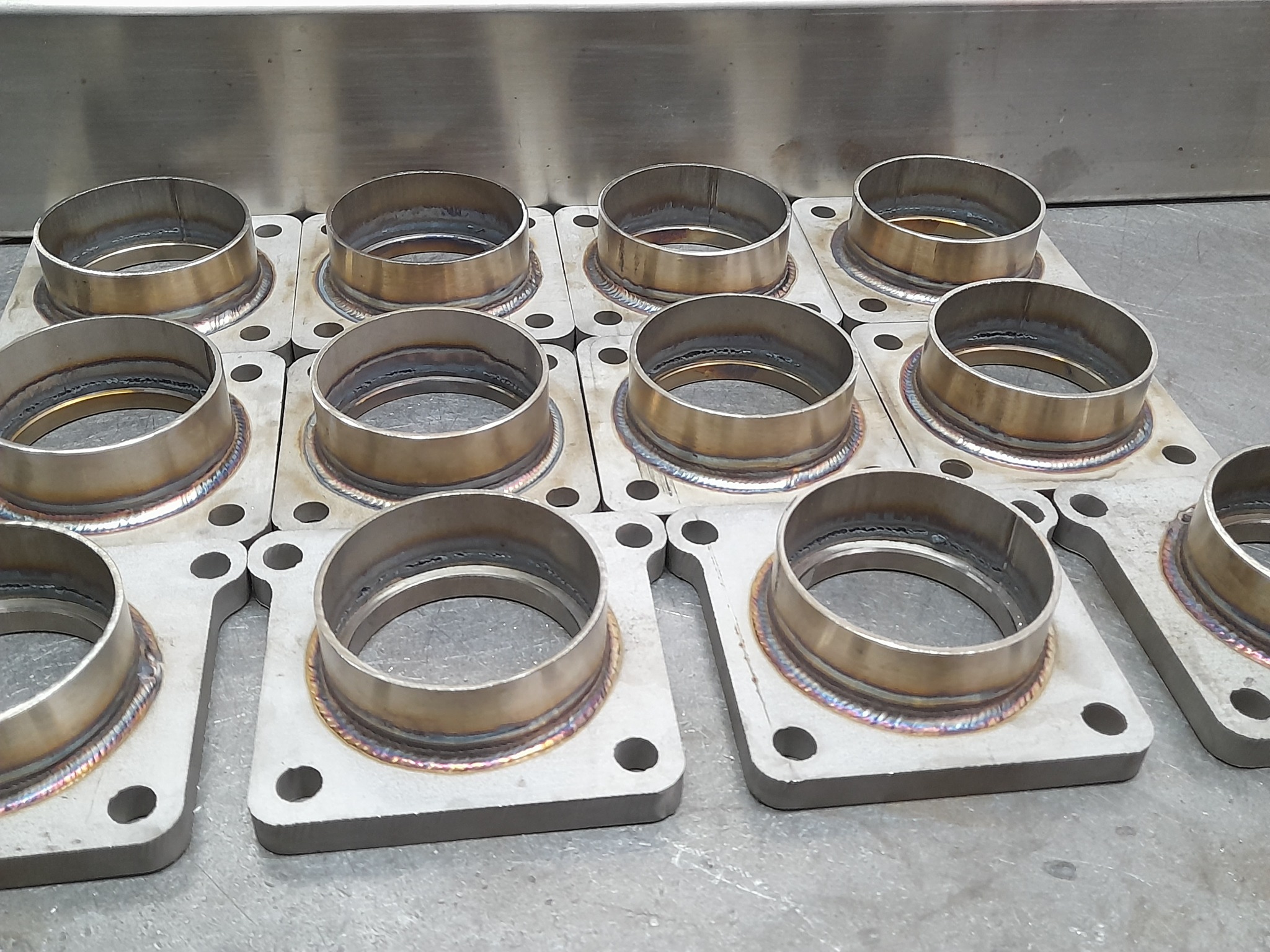 Metalowe elementy z przyspawanymi pierścieniami, ułożone na metalowej powierzchni. Widoczne ślady spawania i otwory montażowe.