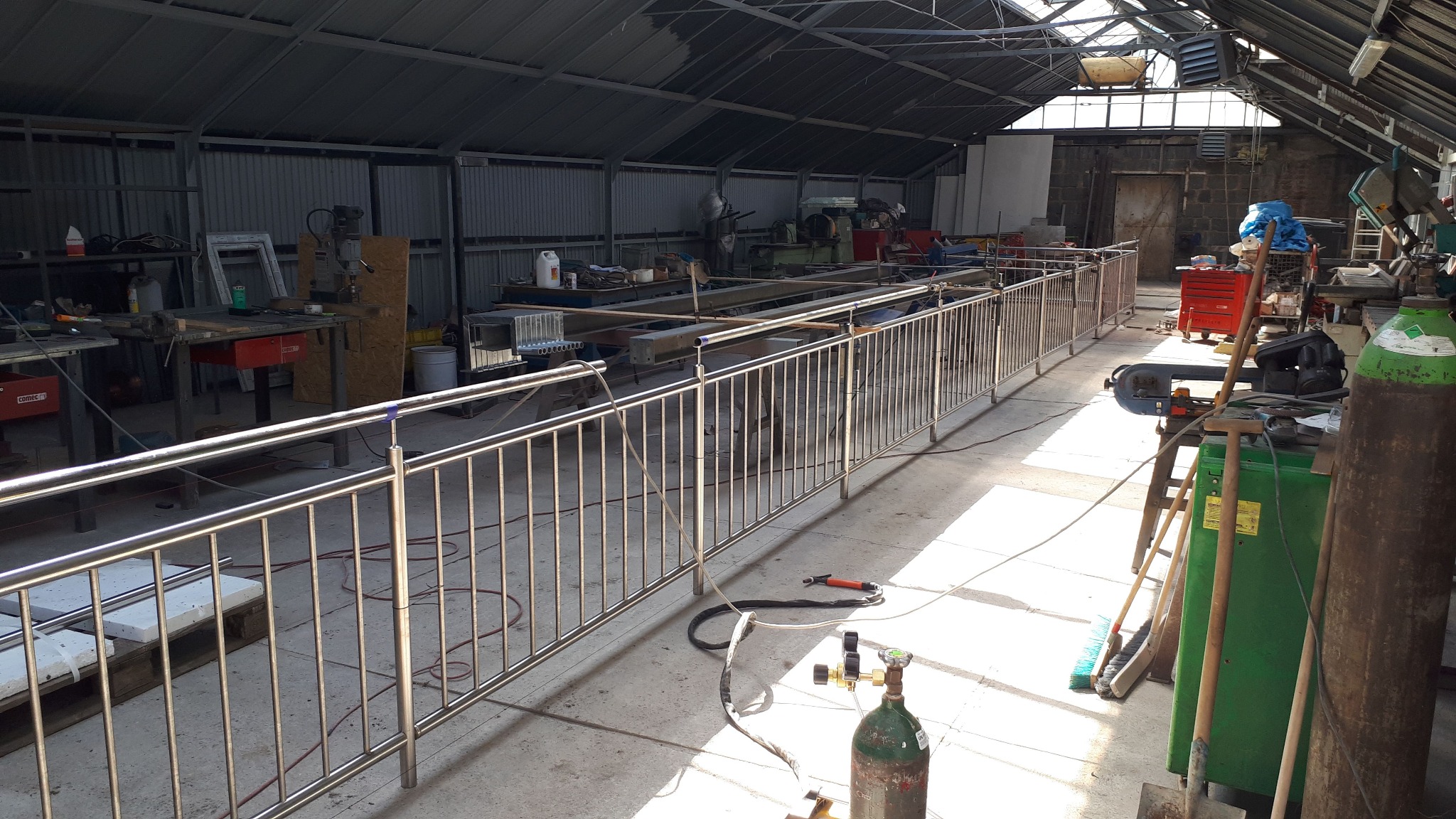 Wnętrze warsztatu z długą, metalową balustradą w trakcie montażu. Widoczne narzędzia spawalnicze, butle z gazem i inne elementy wyposażenia warsztatu.