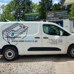 Auto Color Shift - Biały samochód dostawczy z grafiką akumulatora i adresem www.auto-bateria.pl. Reklama firmy na pojeździe, widoczne budynki z reklamami w tle.