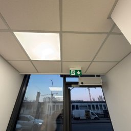 PK PIOSIMPLE SPÓŁKA Z OGRANICZONĄ ODPOWIEDZIALNOŚCIĄ - Kasetonowy sufit podwieszany z oświetleniem LED i systemem wentylacji nad drzwiami z oznaczeniem drogi ewakuacyjnej w nowoczesnym biurze.