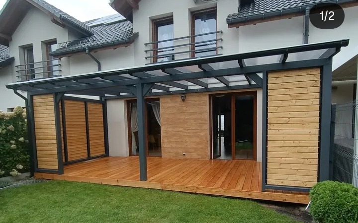 Nowoczesna pergola z drewnianym tarasem przylegająca do domu. Konstrukcja z szarymi słupami i drewnianymi panelami bocznymi. Widok na zadbany trawnik i elewację budynku.