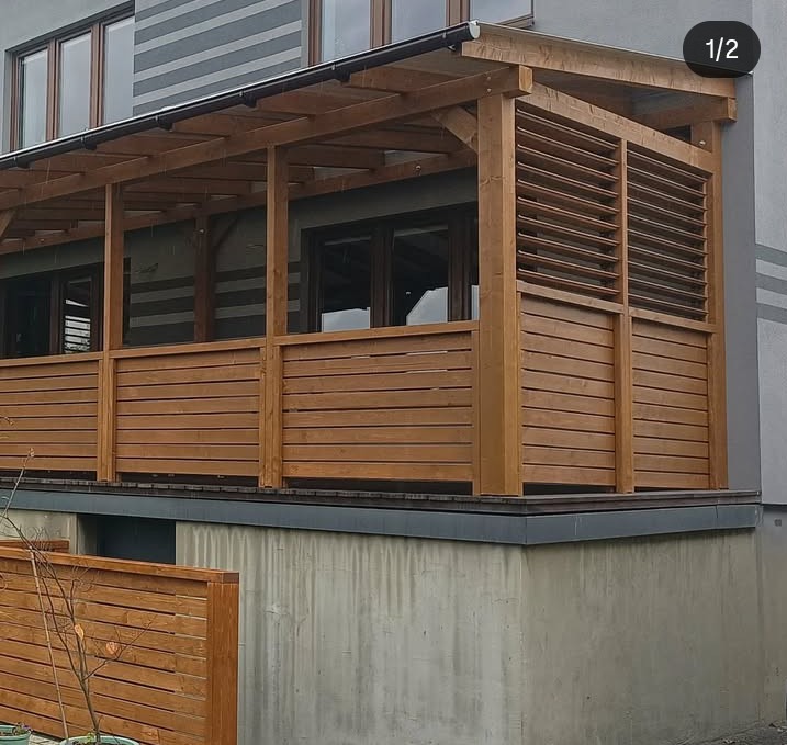 Drewniana pergola z poziomymi deskami, przylegająca do budynku z szarymi pasami. Widoczne okna budynku i fragment niskiego ogrodzenia.