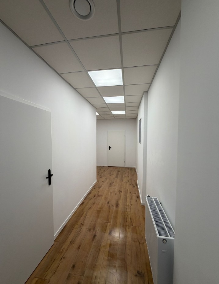 Korytarz biurowy po remoncie: panele podłogowe imitujące drewno, białe ściany, sufit kasetonowy z oświetleniem. Widoczny grzejnik i drzwi z czarną klamką.