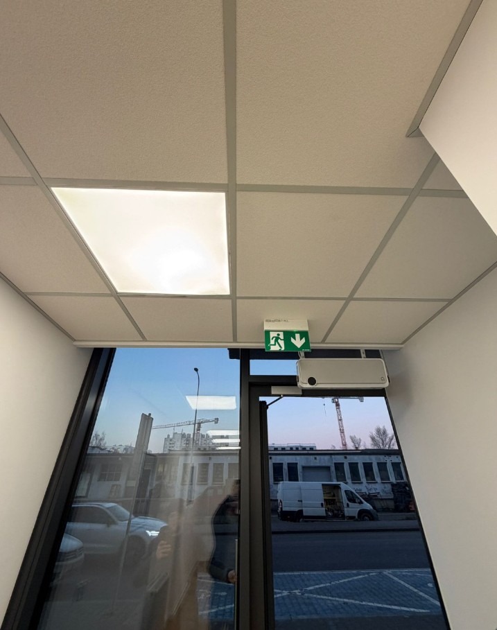 Kasetonowy sufit podwieszany z oświetleniem LED i systemem wentylacji nad drzwiami z oznaczeniem drogi ewakuacyjnej w nowoczesnym biurze.