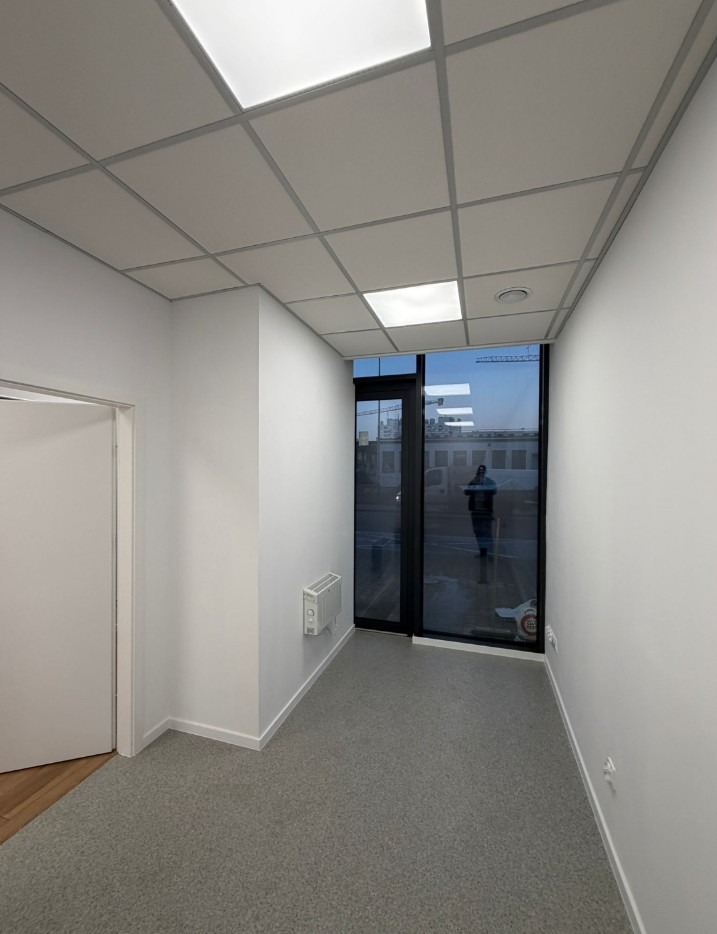 Wnętrze biura po remoncie w Orzesze: białe ściany, sufit podwieszany kasetonowy, szara podłoga, okno z widokiem na zewnątrz. Minimalistyczny design.