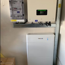 PanSolarnypl - Biała bateria i inwerter fotowoltaiczny SOFAR zamontowane na ścianie, obok skrzynka z bezpiecznikami. Widoczne logo PanSolarny.pl.