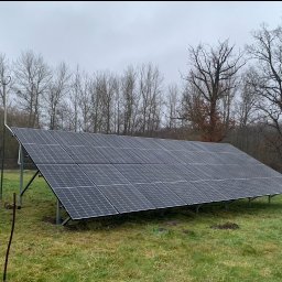 PanSolarnypl - Panel fotowoltaiczny na trawiastym terenie, z widocznymi drzewami w tle i pochmurnym niebem. Widoczny wózek z kartonami. Instalacja naziemna.