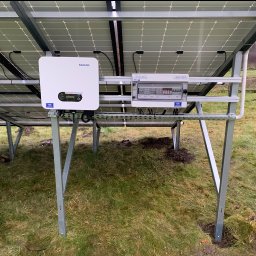 PanSolarnypl - Montaż paneli fotowoltaicznych z widocznym inwerterem Sofar i skrzynką bezpiecznikową na metalowej konstrukcji, na tle trawy. Widok od spodu instalacji.