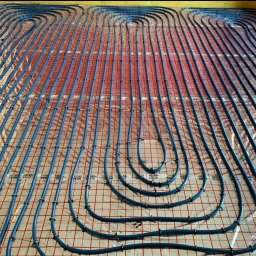 PanSolarnypl - Niebieskie rury ogrzewania podłogowego ułożone równolegle na folii z czerwoną siatką, zabezpieczone czarnymi klipsami. Widok z góry, instalacja przed zalaniem posadzki.