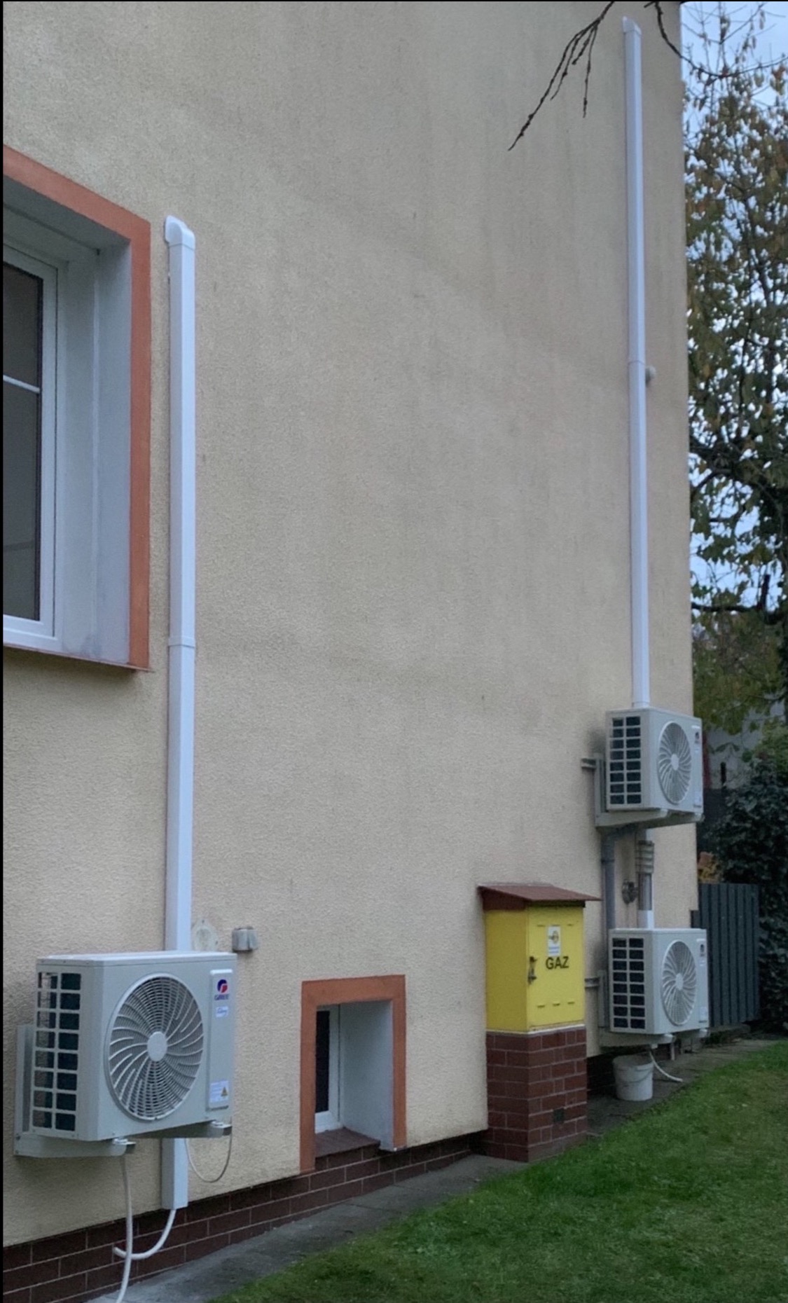 Elewacja budynku z zamontowanymi jednostkami zewnętrznymi klimatyzacji, rurami odprowadzającymi i skrzynką gazową. Widoczne okno i fragment trawnika.