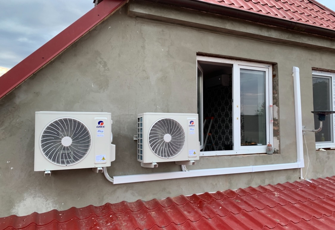 Dwa klimatyzatory marki Gree zamontowane na szarej ścianie budynku z czerwonym dachem, widoczne okna z białymi ramami i białe listwy maskujące instalację.