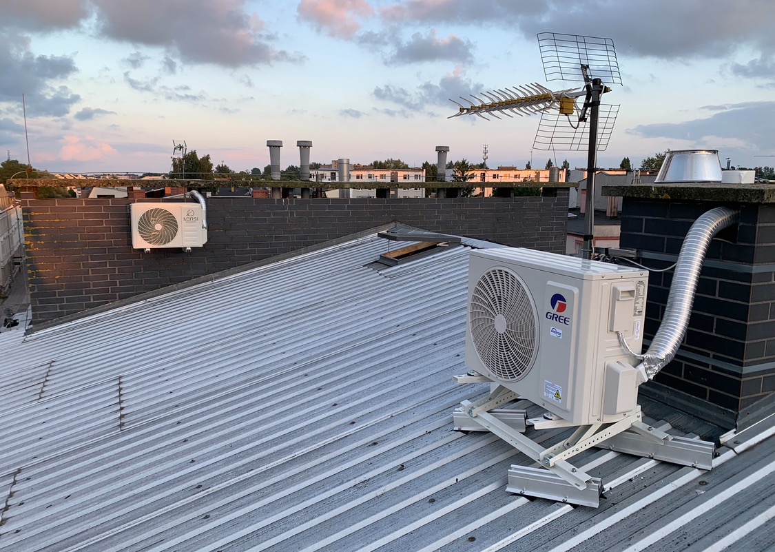 Klimatyzator GREE zamontowany na dachu z blachy falistej, widoczny komin i antena. W tle zabudowa miejska i zachmurzone niebo o zmierzchu. Drugi klimatyzator na ścianie budynku.