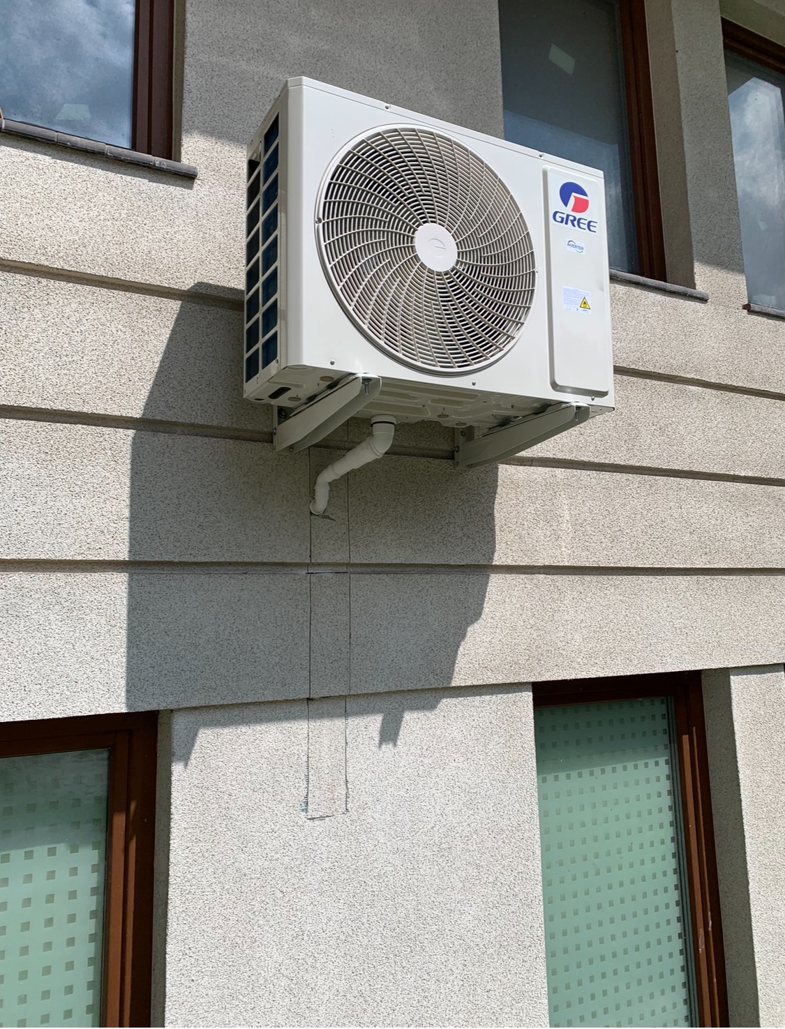 Biały klimatyzator marki Gree zamontowany na elewacji budynku z widocznymi oknami i cieniem. Widoczne rurki odprowadzające skropliny.