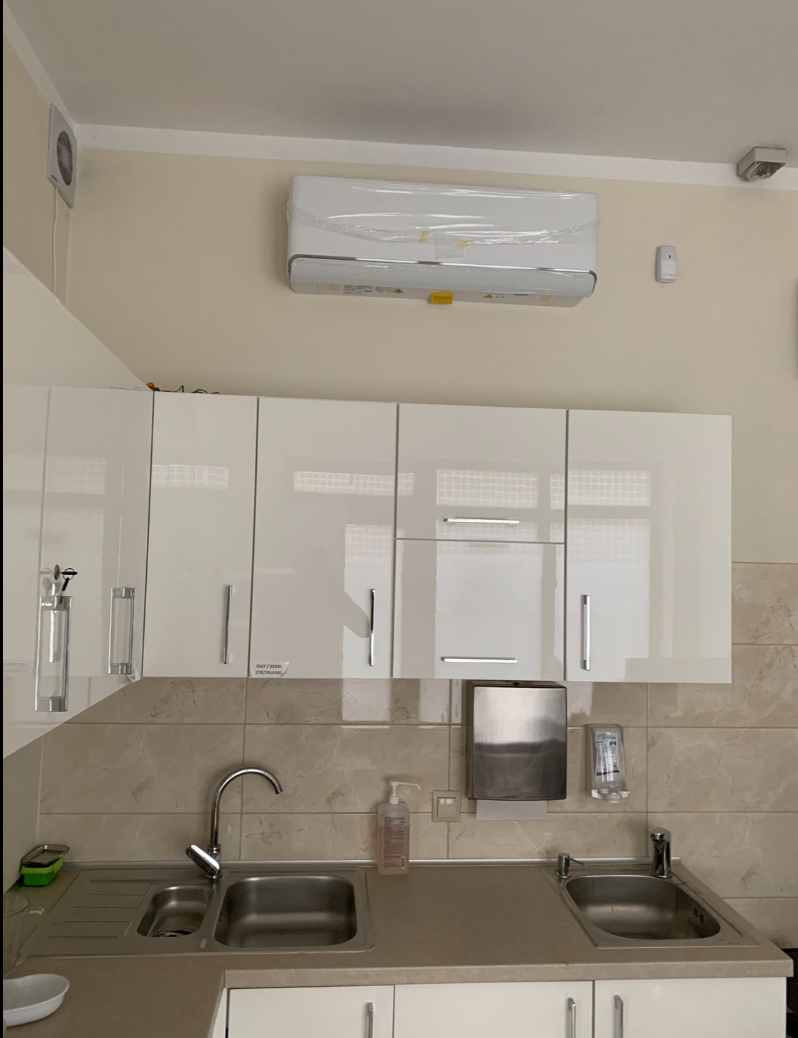 Biała klimatyzacja na ścianie nad umywalką z baterią i szafkami. Widoczny dozownik mydła i ręczników. Nowoczesne wnętrze z płytkami imitującymi kamień.