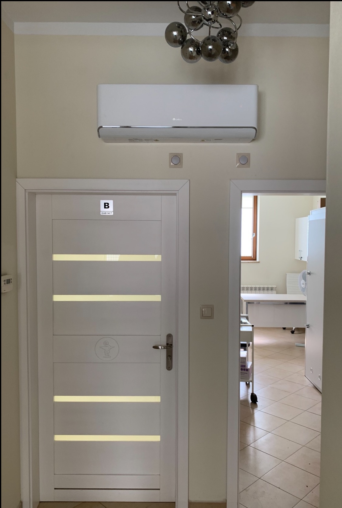 Biały klimatyzator ścienny nad drzwiami do gabinetu z widokiem na wnętrze. Nowoczesne oświetlenie sufitowe. Jasne ściany i płytki podłogowe.