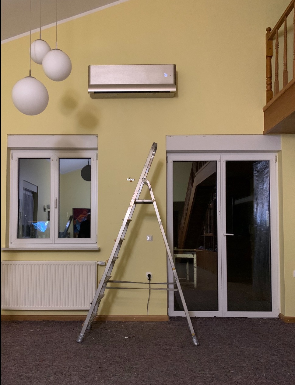 Klimatyzator ścienny w kolorze szampańskim zamontowany nad oknem, z drabiną opartą o ścianę. Widoczne lampy wiszące i fragment balustrady na antresoli.