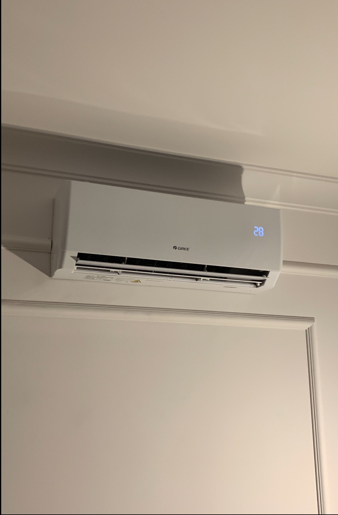 Biały klimatyzator marki Gree zamontowany na ścianie z dekoracyjną listwą. Na urządzeniu widoczny wyświetlacz z temperaturą 28 stopni.