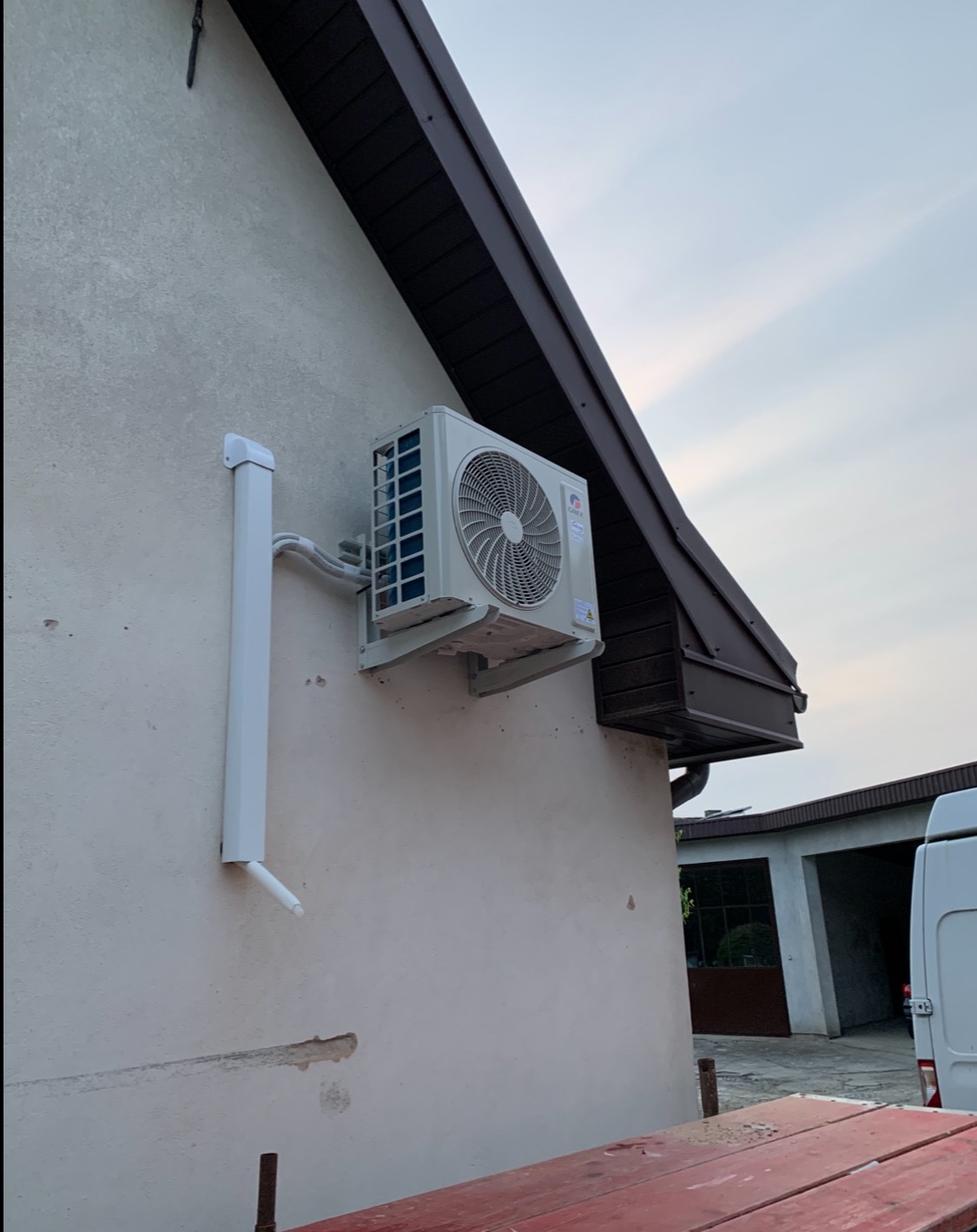 Biały klimatyzator zamontowany na elewacji domu z brązowym dachem, widoczna rynna i fragment budynku gospodarczego w tle, ujęcie z dołu.