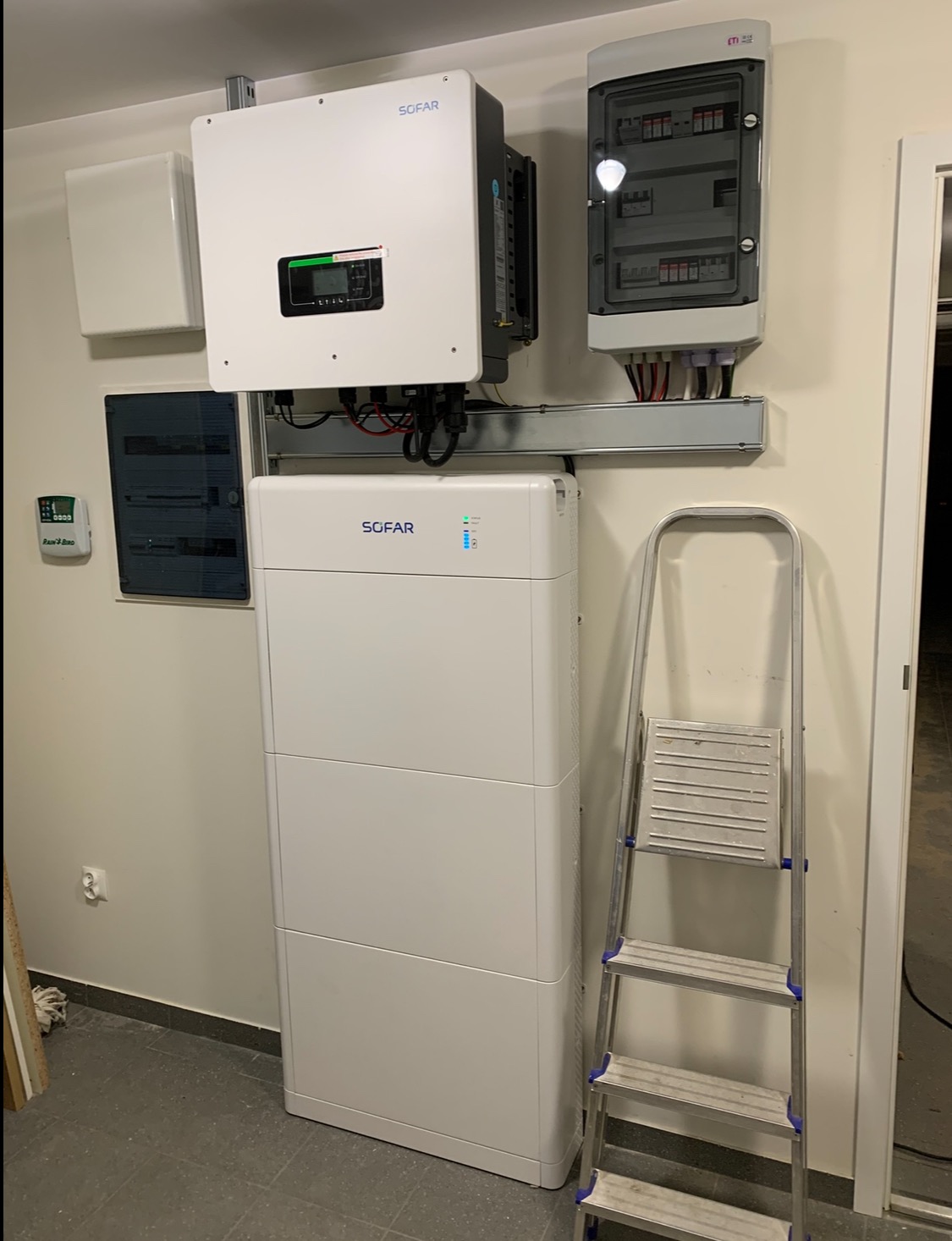 Biała bateria i inwerter Sofar zamontowane na ścianie w pomieszczeniu, obok skrzynka elektryczna i drabina. Instalacja fotowoltaiczna wewnątrz budynku.