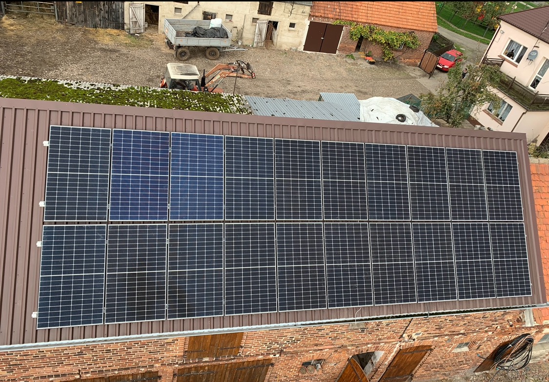 Panel słoneczny na brązowym dachu budynku gospodarczego z cegły, widok z góry. Część paneli ma inny odcień. W tle zabudowania gospodarcze i traktor.