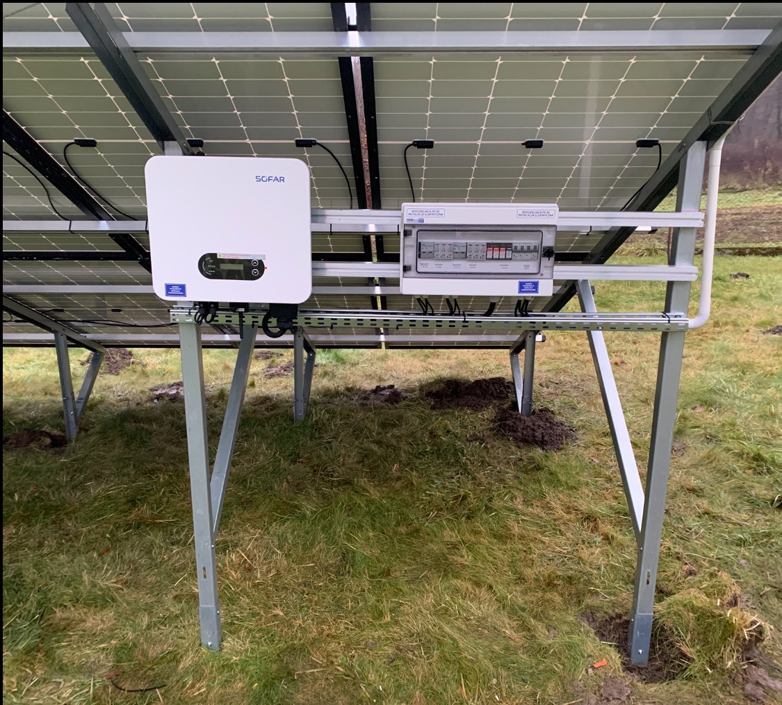 Montaż paneli fotowoltaicznych z widocznym inwerterem Sofar i skrzynką bezpiecznikową na metalowej konstrukcji, na tle trawy. Widok od spodu instalacji.