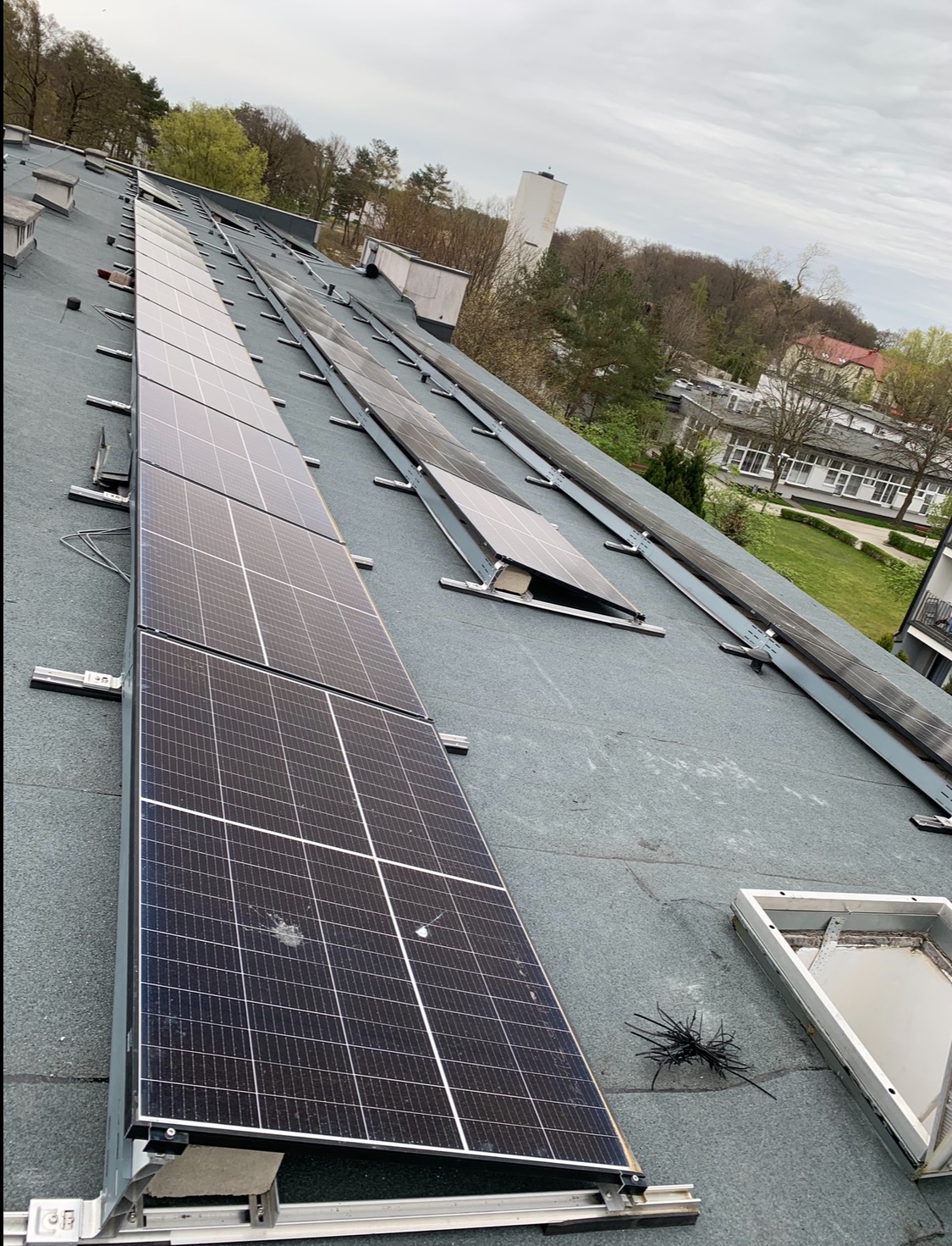 Rząd paneli fotowoltaicznych na płaskim dachu budynku, widok z góry, pochmurne niebo. Widoczne elementy montażowe i fragmenty instalacji.