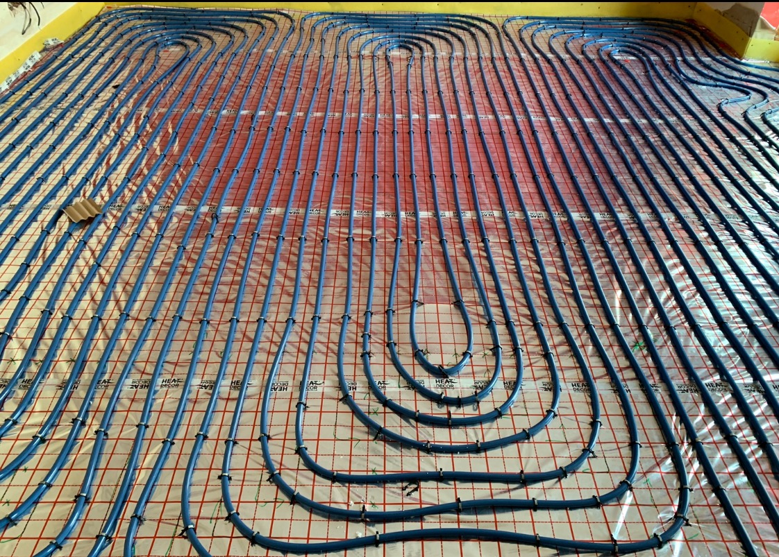 Niebieskie rury ogrzewania podłogowego ułożone równolegle na folii z czerwoną siatką, zabezpieczone czarnymi klipsami. Widok z góry, instalacja przed zalaniem posadzki.