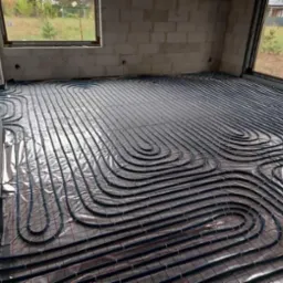 Montaż rur ogrzewania podłogowego na folii z siatką w surowym wnętrzu budynku w stanie deweloperskim, widoczne okna i ściany z betonu komórkowego.