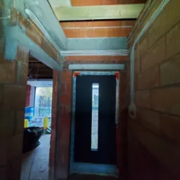 Wnętrze budynku w trakcie budowy, widoczne przewody elektryczne ułożone wzdłuż ściany z cegły ceramicznej, zamontowane drzwi wejściowe z długim przeszkleniem.