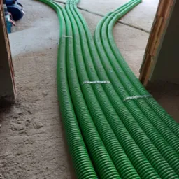 Wiązka zielonych, karbowanych rur wentylacyjnych ułożona na surowym betonowym podłożu w trakcie montażu, widoczne metalowe opaski mocujące i fragmenty ścian w stanie surowym.