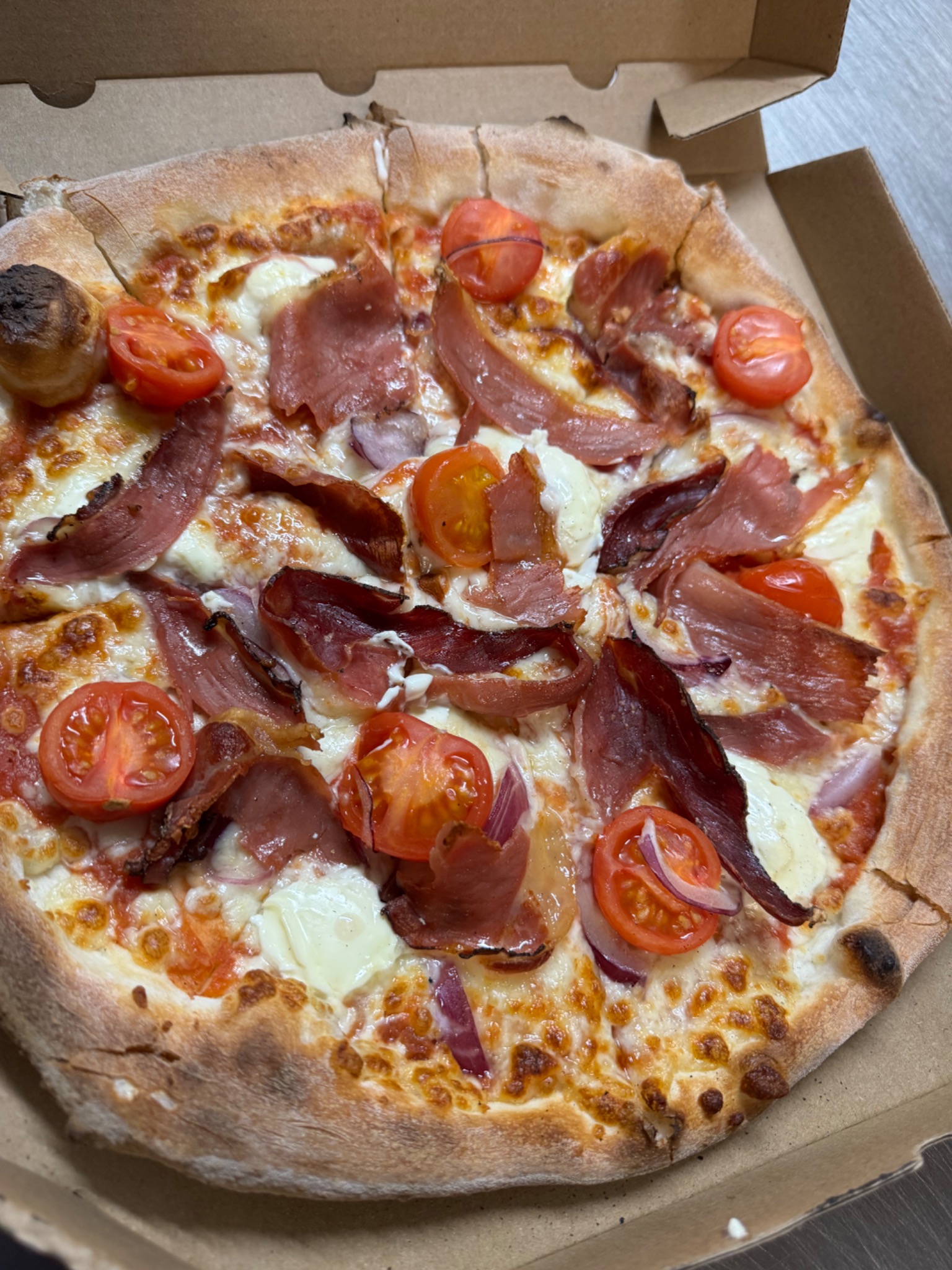 Pizza z szynką parmeńską, pomidorkami cherry i czerwoną cebulą w kartonowym pudełku. Widoczne przypalone brzegi ciasta i świeże składniki.