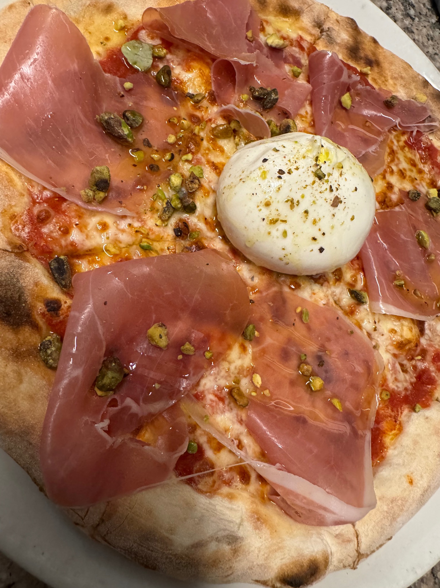 Pizza z burratą, szynką parmeńską i pistacjami na cienkim cieście, z widocznymi przypaleniami na brzegach. Ujęcie z bliska, podkreślające świeżość składników.