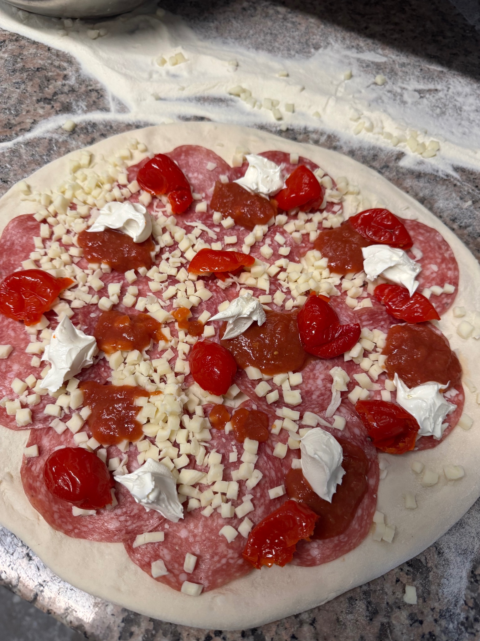 Pizza w trakcie przygotowywania: salami, ser mozzarella w kostce, pomidory koktajlowe i sos na surowym cieście. Widoczny blat posypany mąką.